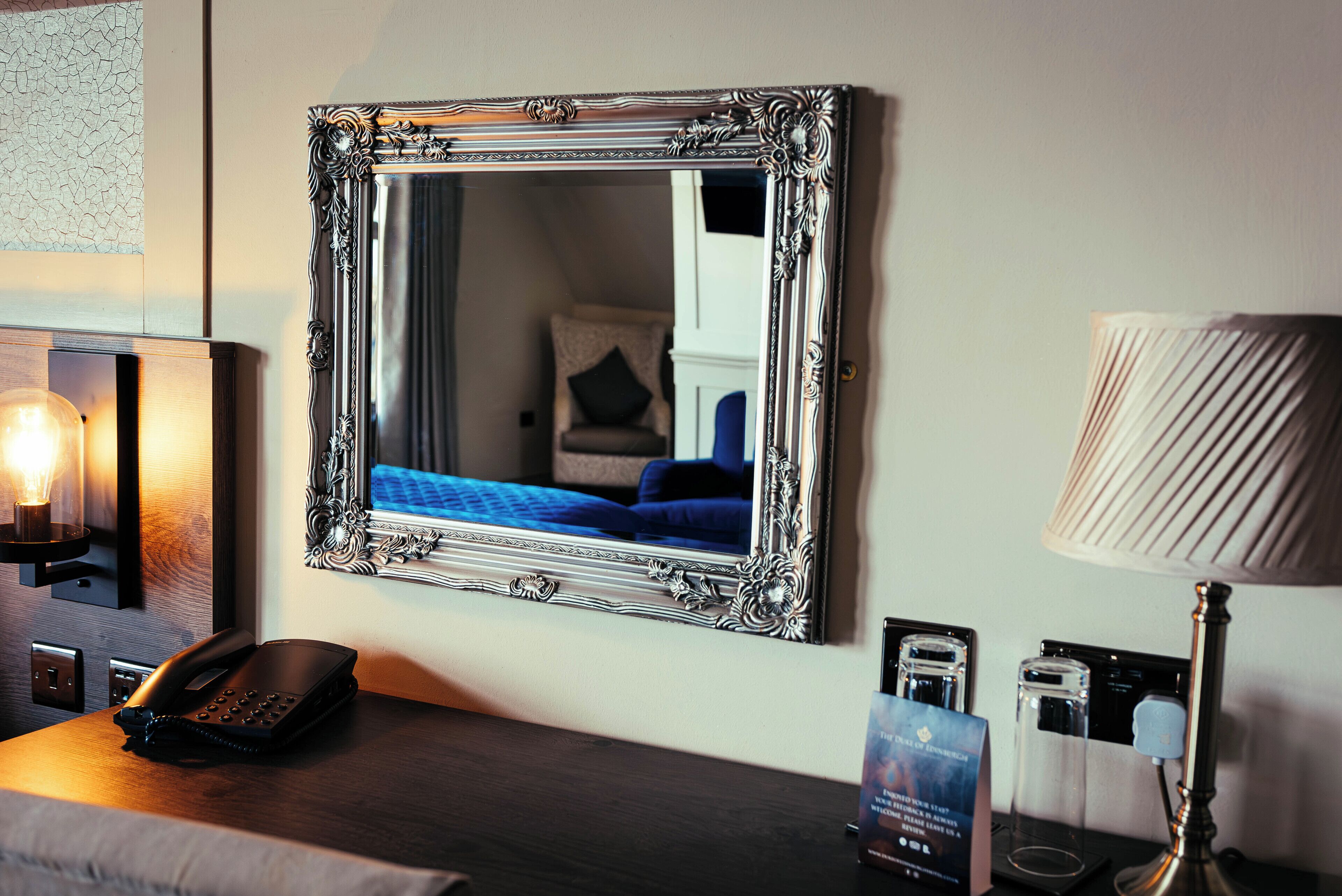 Una camera d'hotel con un ampio specchio decorato, un telefono sul comodino, una lampada e un quadro appeso alla parete.