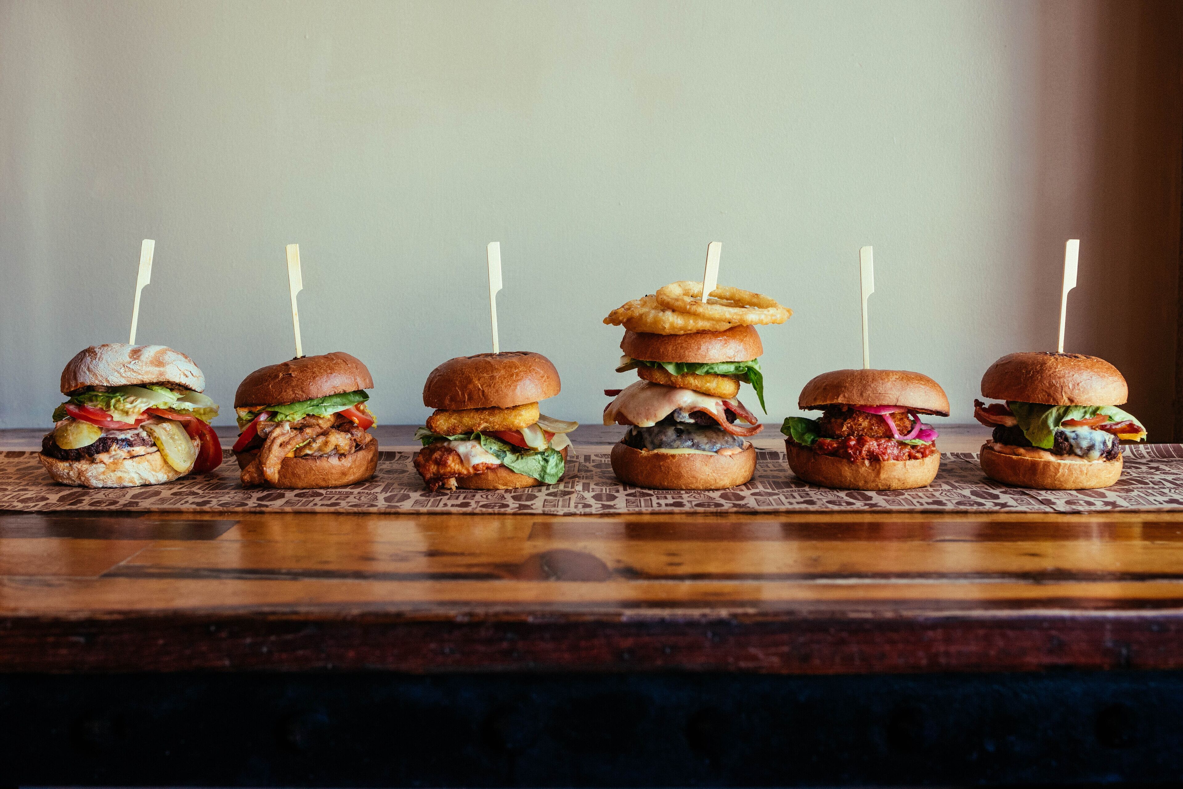 Seis hamburguesas con palillos de dientes, dispuestas sobre una mesa de madera frente a una pared lisa.