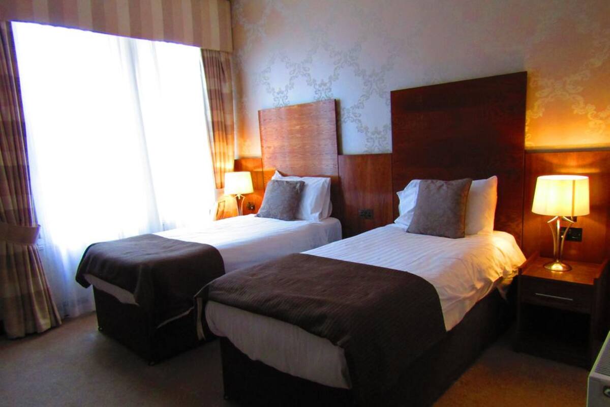 A hotel room with two beds, a large window with curtains, and a nightstand with a lamp.