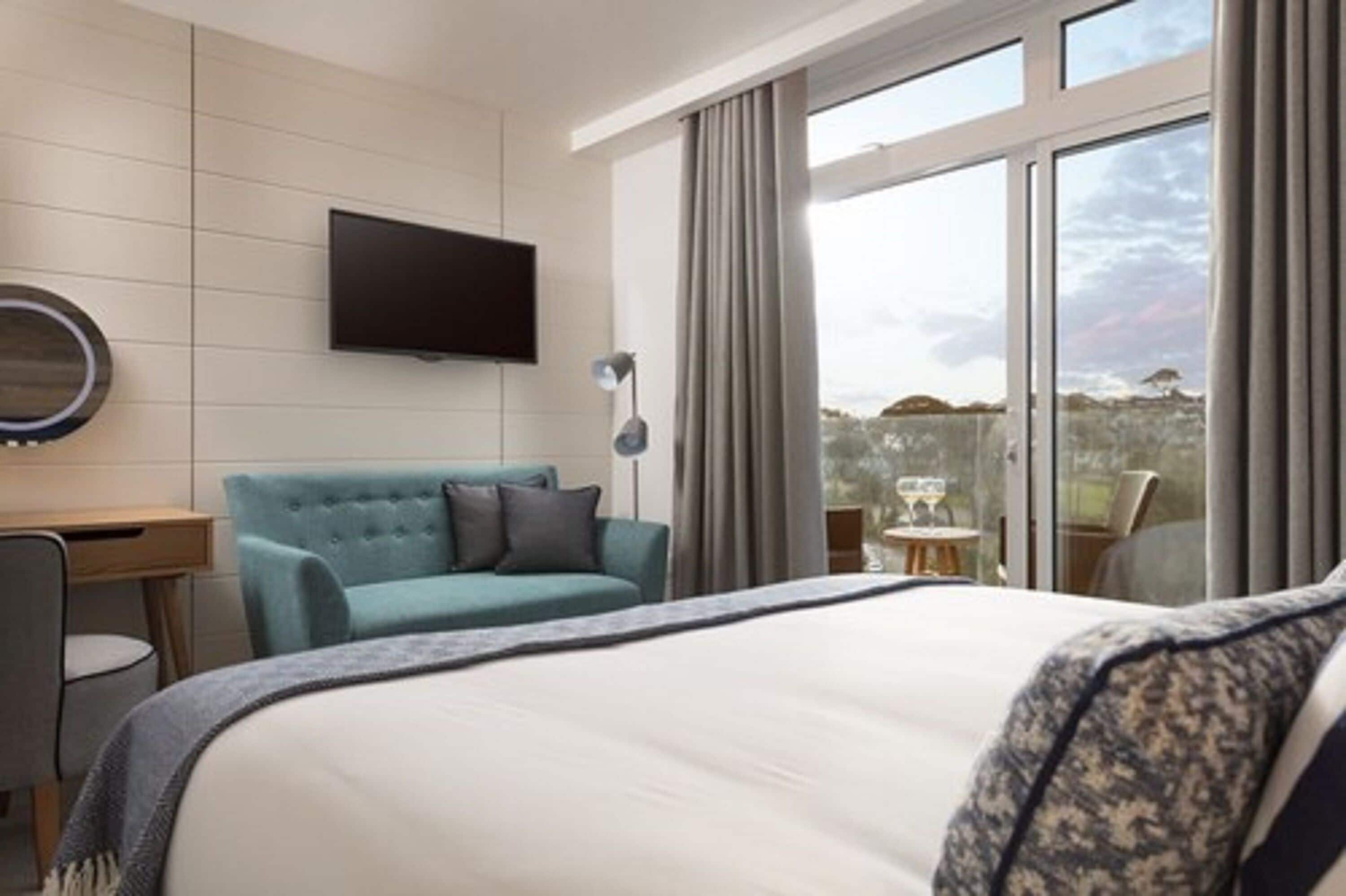 Hotelkamer met een bed, een blauwe bank, een bureau, een flatscreen-tv en een groot raam met gordijnen.