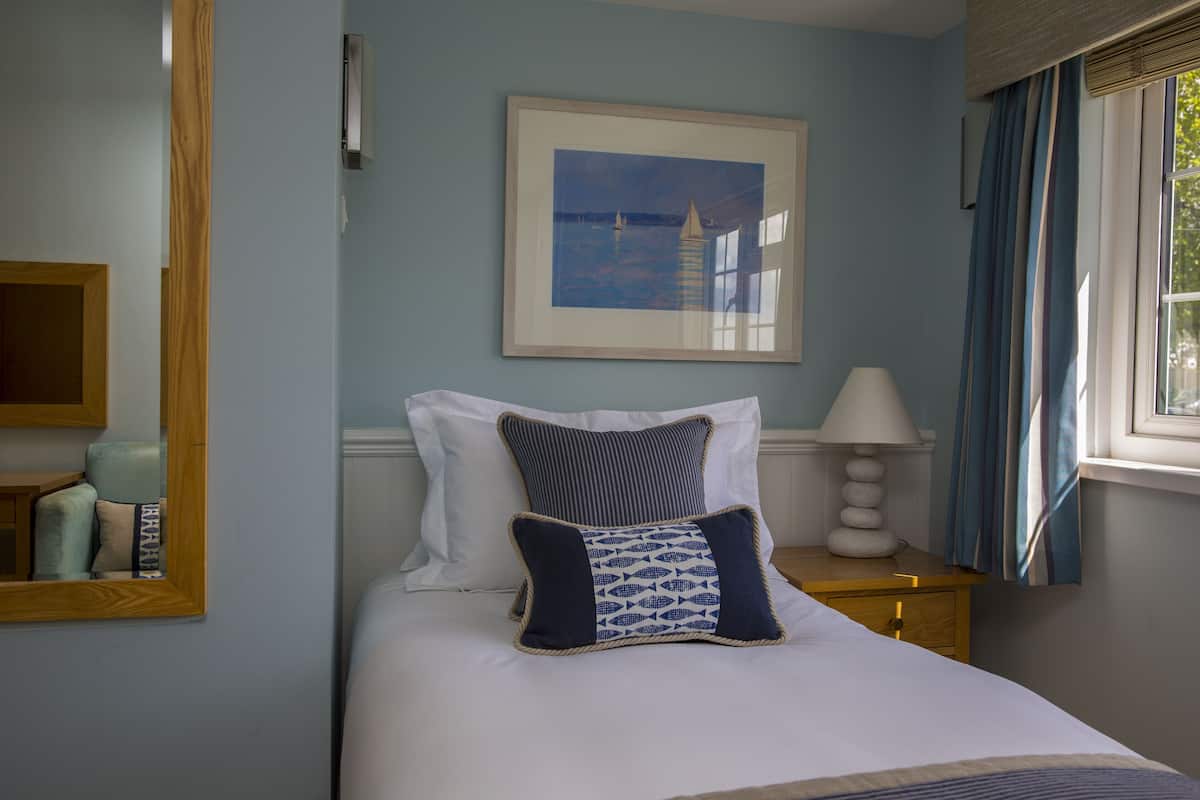 A neatly arranged bedroom with a bed, pillows, a bedside table with a lamp, a framed picture on the wall, and a window with curtains.