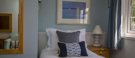 A neatly arranged bedroom with a bed, pillows, a bedside table with a lamp, a framed picture on the wall, and a window with curtains.