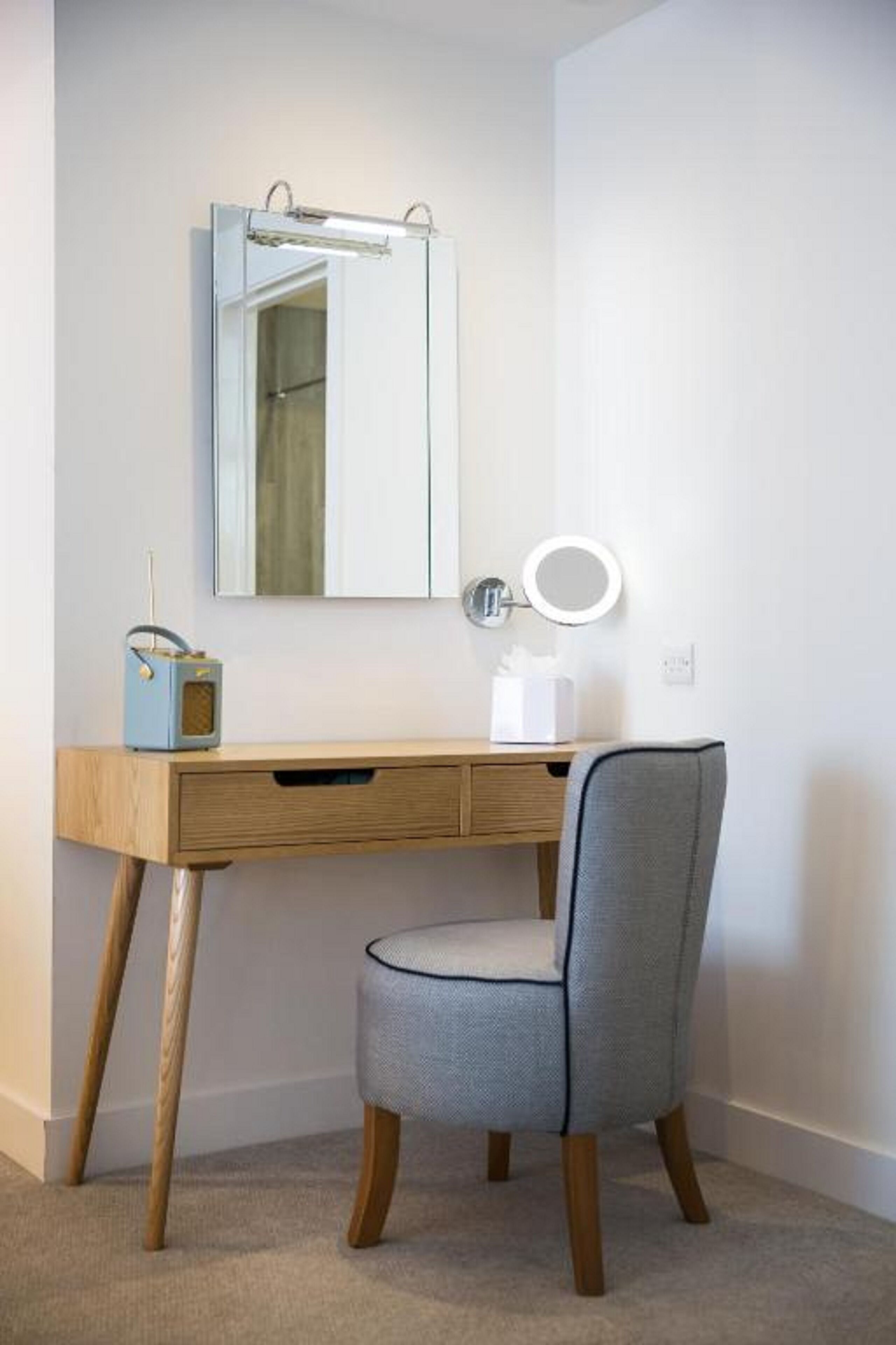 A small, modern bathroom with a wooden vanity, a mirror, a round vanity light, and a chair.