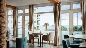 Un comedor con vista al mar, con una mesa dispuesta con vasos y una carta, y sillas con tapicería de tela clara.