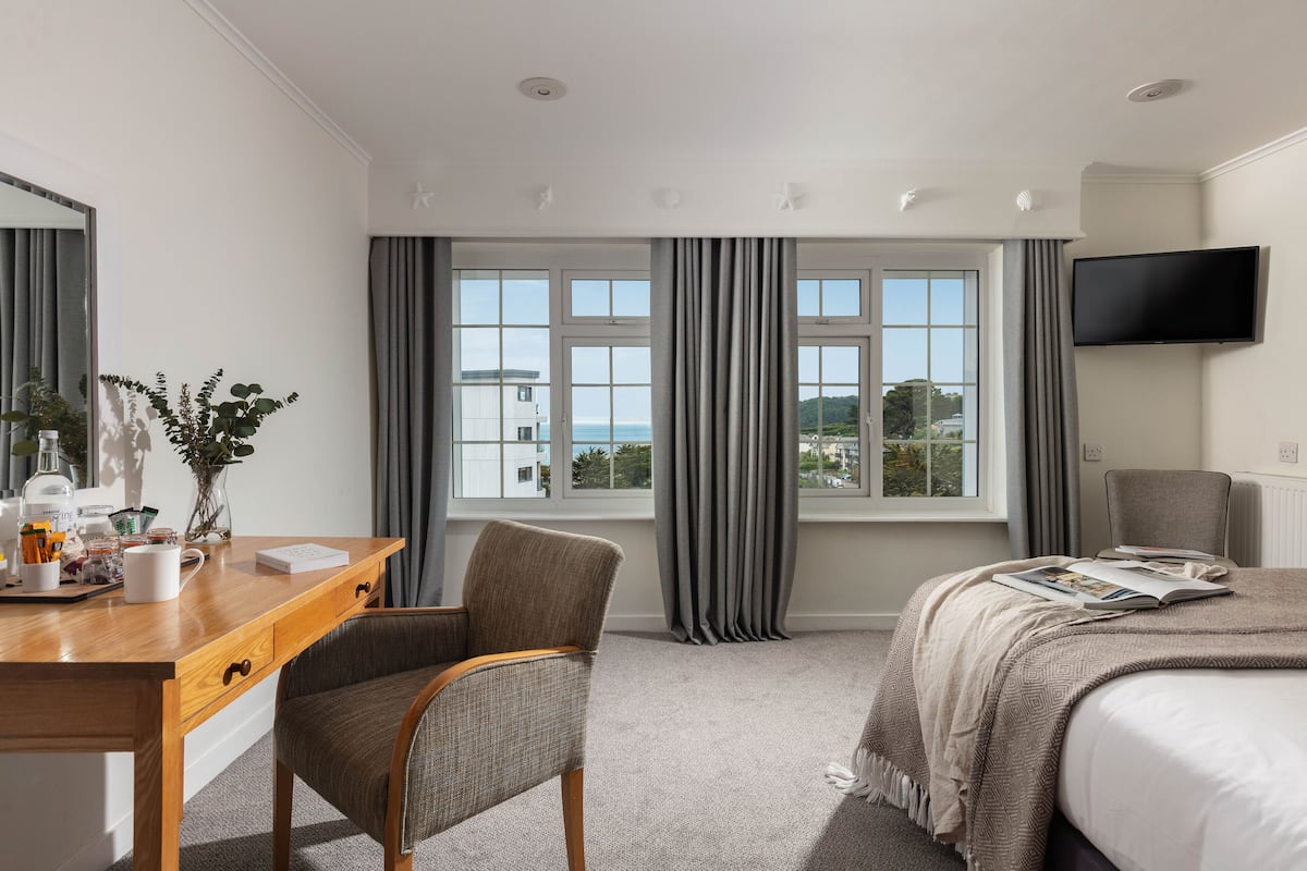 A hotel room with a wooden desk, a chair, a bed with a beige blanket, a TV mounted on the wall, and a window with curtains.