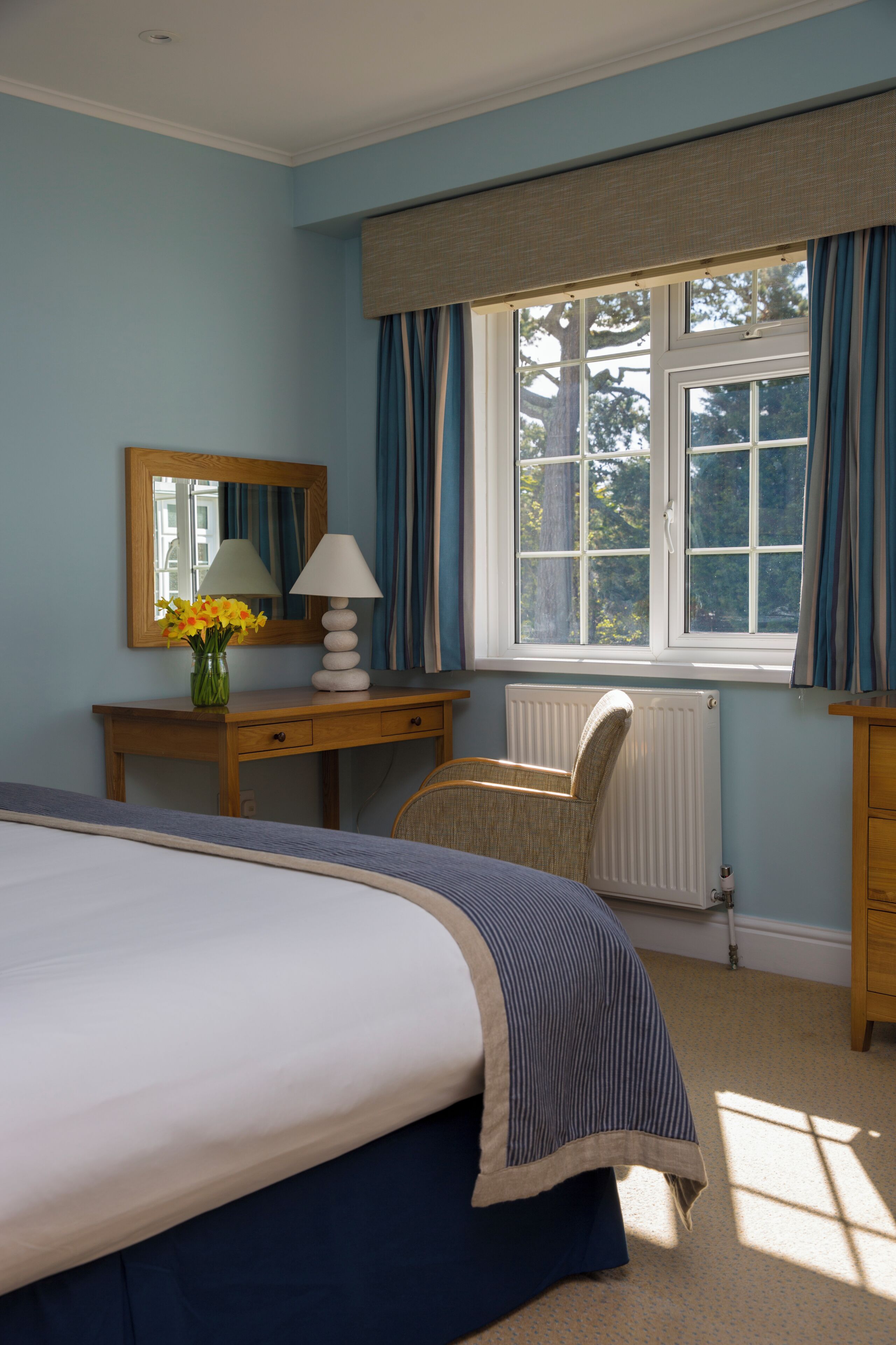 A bedroom with a bed, a desk with a lamp, a chair, a dresser, a radiator, and a window with curtains.