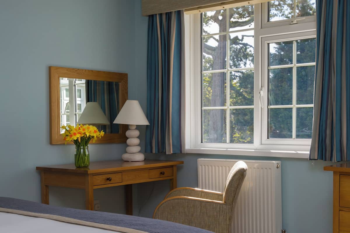 A bedroom with a bed, a desk with a lamp, a chair, a dresser, a radiator, and a window with curtains.
