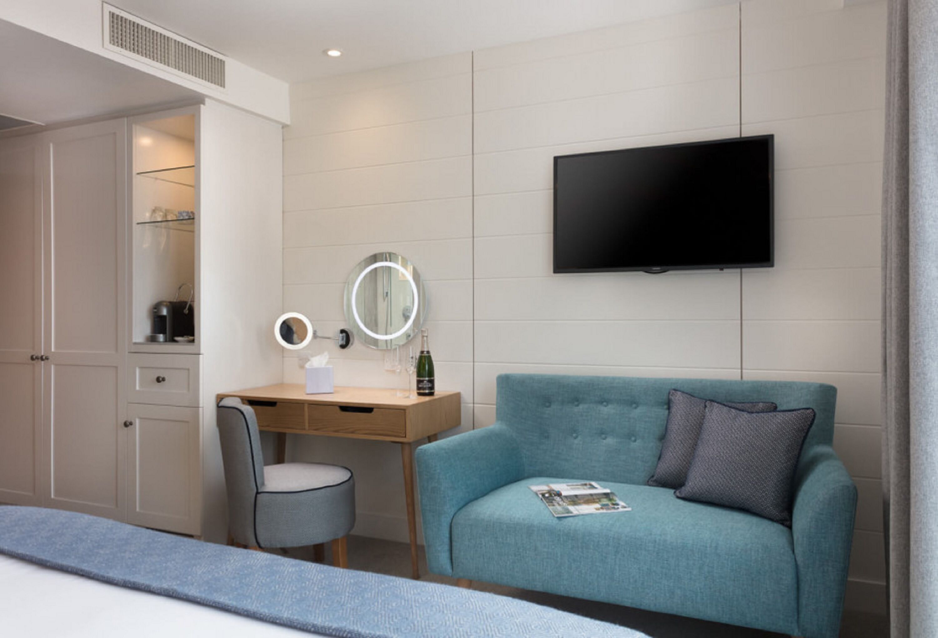 A modern hotel room with a bed, a desk with a chair, a blue sofa, a television, and a wardrobe.