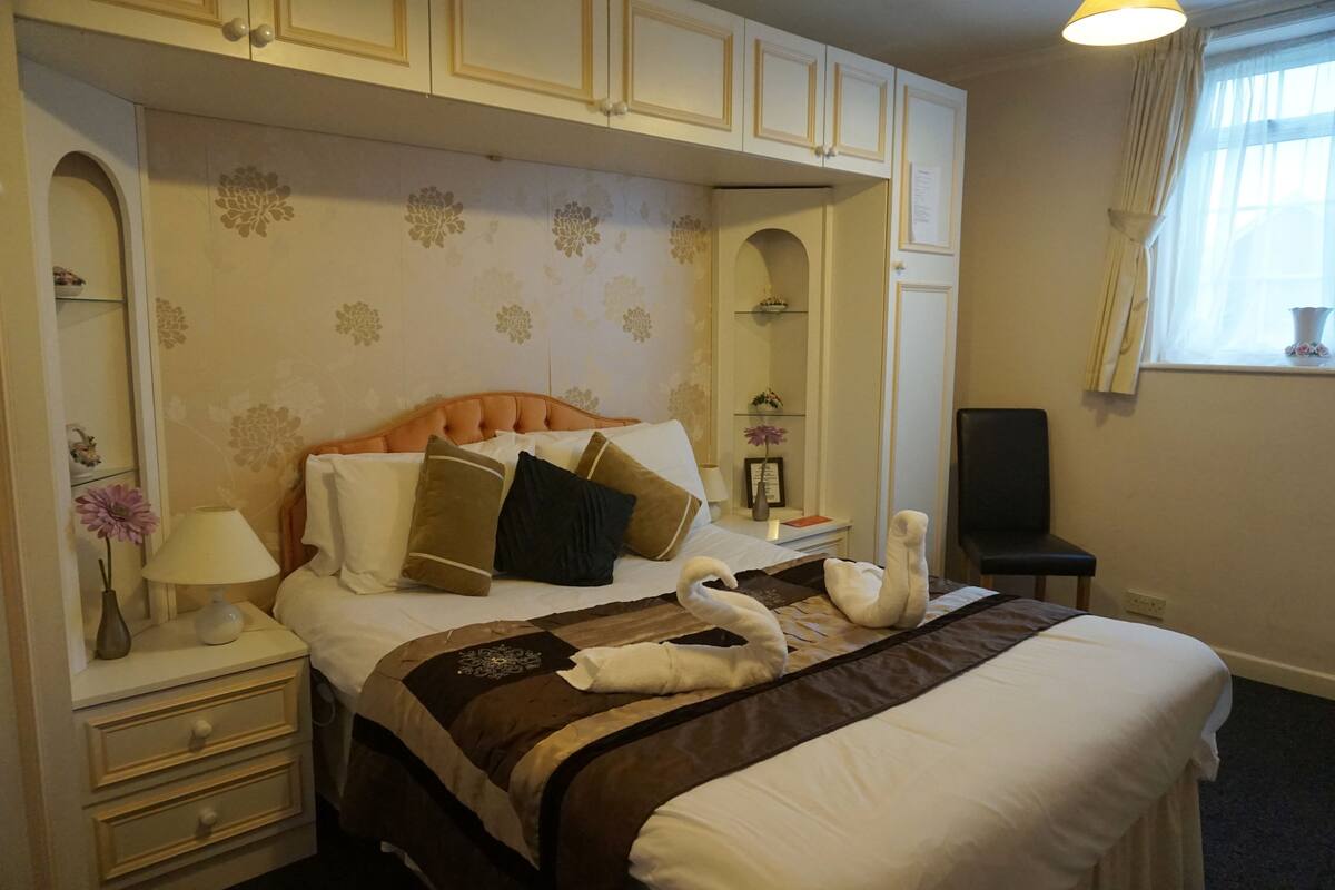 A hotel room with a bed, a nightstand, a lamp, a vase with flowers, a chair, and a window with curtains.