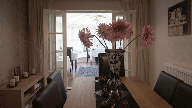 A dining area with a wooden table, black chairs, and a vase with pink flowers. There is a window with curtains and a painting on the wall.