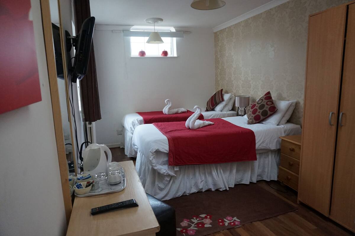 A hotel room with two beds, each with a red blanket and white pillows, a wooden wardrobe, a desk with a kettle and glasses, and a window with a white curtain.