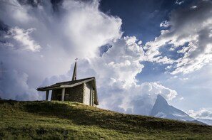 A small stone chapel with a cross on top, surrounded by a grassy hill and a mountain in the background.