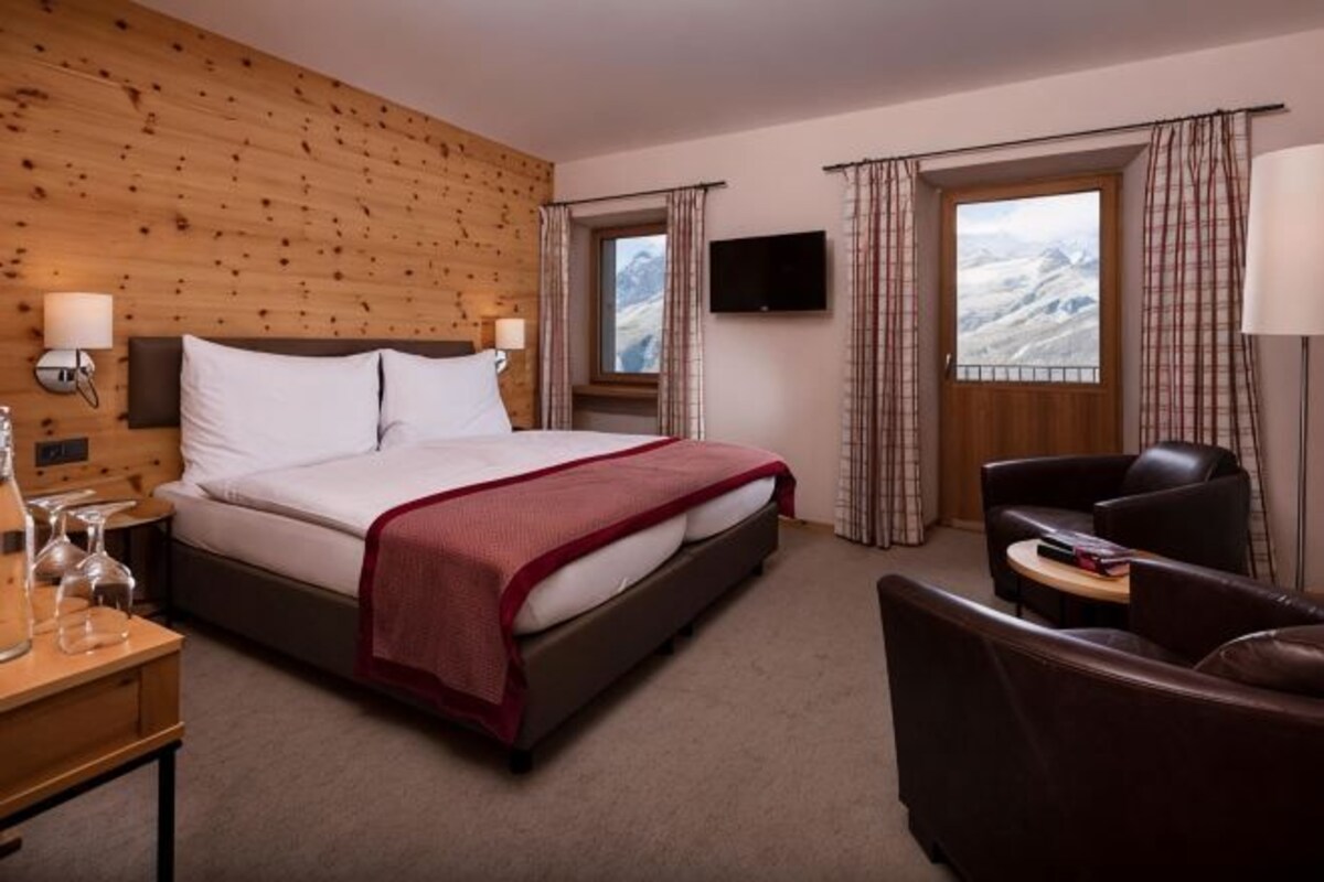 A hotel room with a wooden wall, a bed with white linens, a TV, a window with curtains, and a seating area with a table.