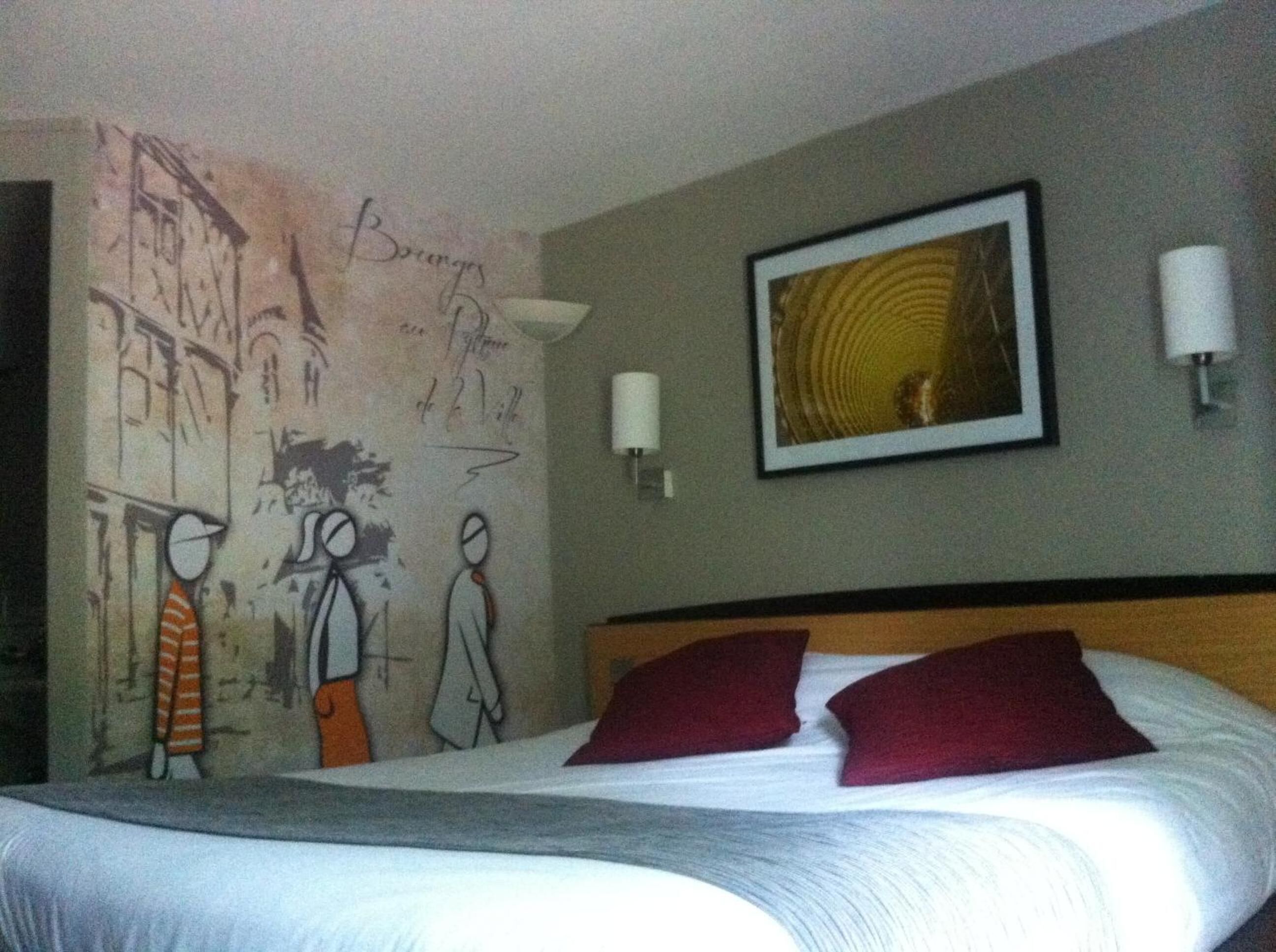 A hotel room with a bed, two pillows, a framed picture on the wall, and a wall with graffiti.