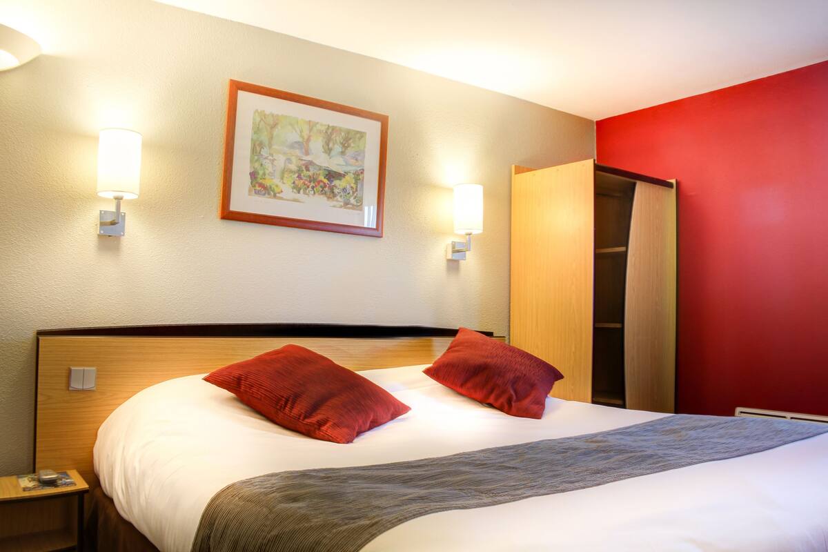 A hotel room with a bed, two red pillows, a framed picture on the wall, and a nightstand with a lamp.