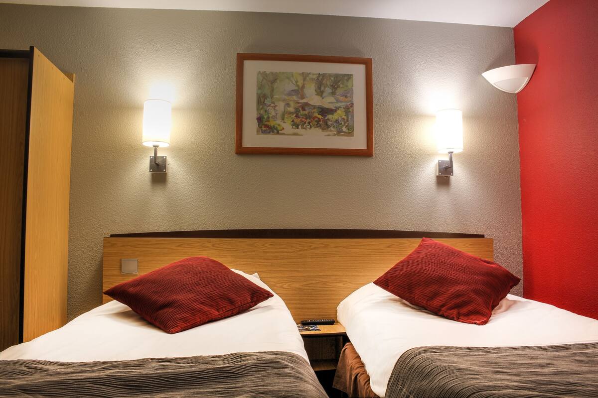 A hotel room with two beds, a wooden headboard, a framed painting on the wall, and two wall-mounted lamps.