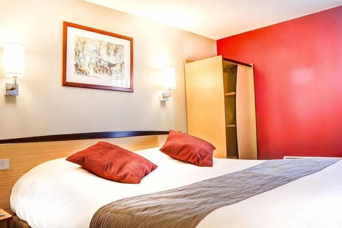 A hotel room with a bed, a red wall, a wooden wardrobe, and a framed picture on the wall.