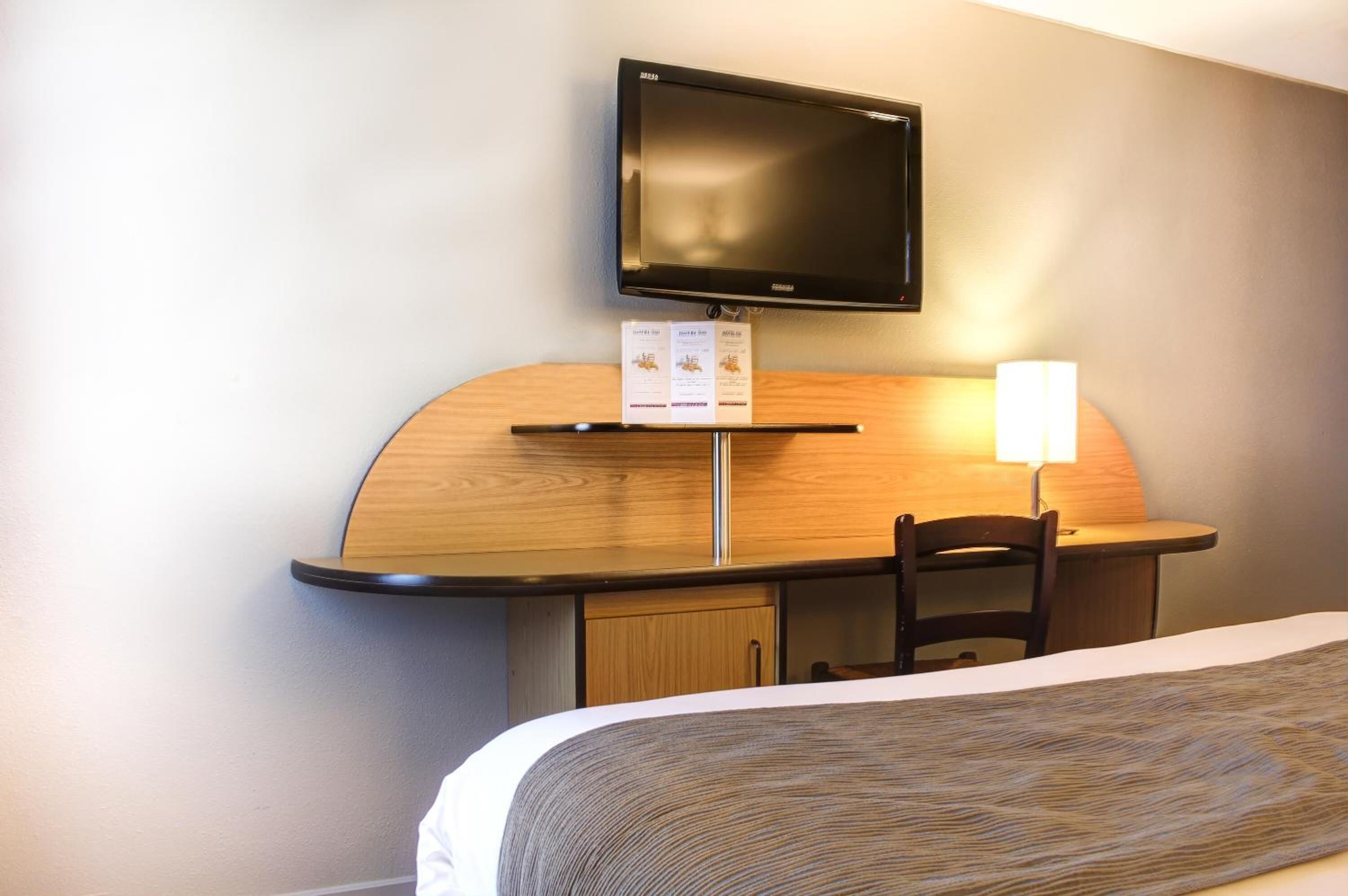 Camera d'albergo con un letto, una scrivania con televisione a schermo piatto, una sedia e una lampada.