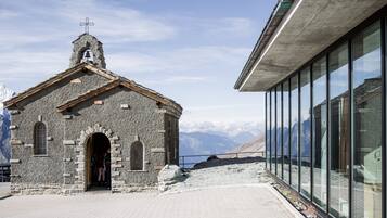 A small stone chapel with a bell tower and arched doorway, adjacent to a modern glass building with a mountain view.