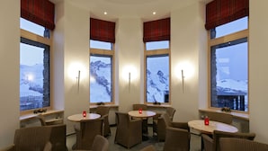 A cozy room with four round tables and wicker chairs, large windows with red plaid curtains, and a view of snow-covered mountains. - 3100 Kulmhotel Gornergrat (Zermatt)