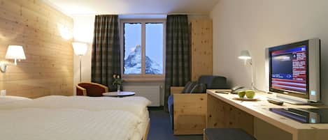 Habitación de hotel con dos camas, un escritorio con computadora y televisor, una silla, una mesita con lámpara y una ventana con cortinas.