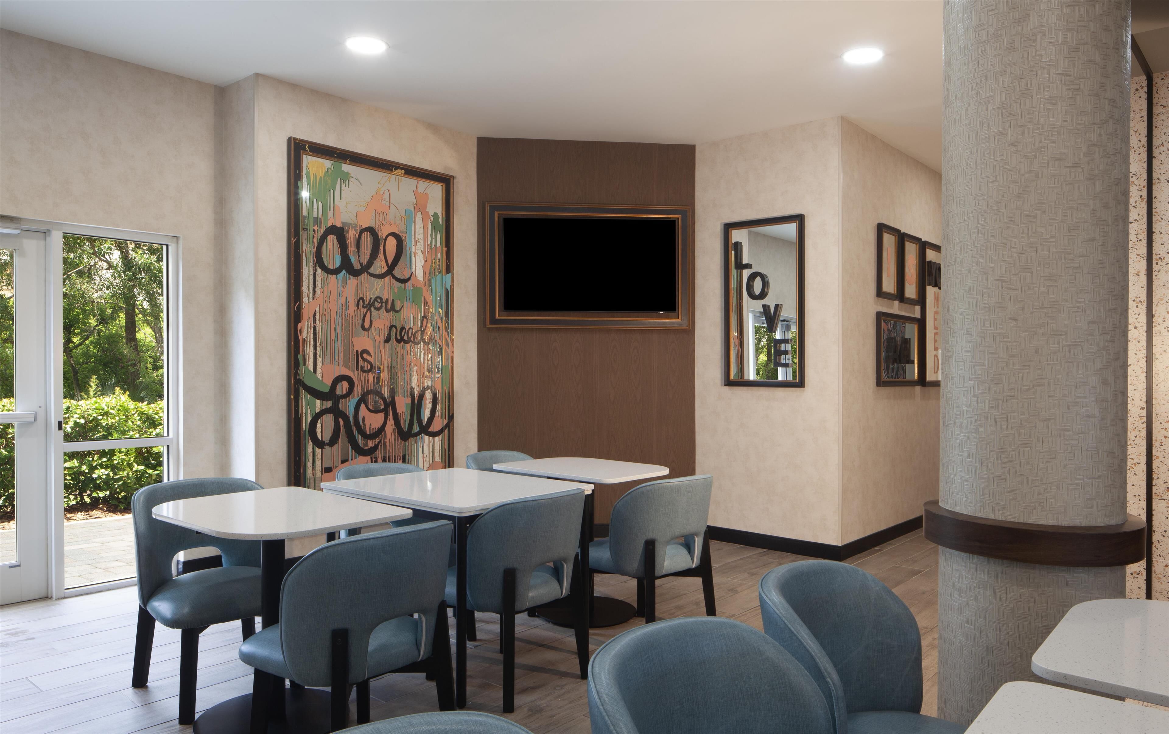 Un comedor moderno con mesas y sillas, un cuadro abstracto grande y un televisor en la pared.