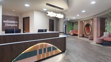 Een moderne receptiebalie met een bord met de tekst 'Welcome to Hampton', een receptie en een zithoek met comfortabele stoelen en poefs.