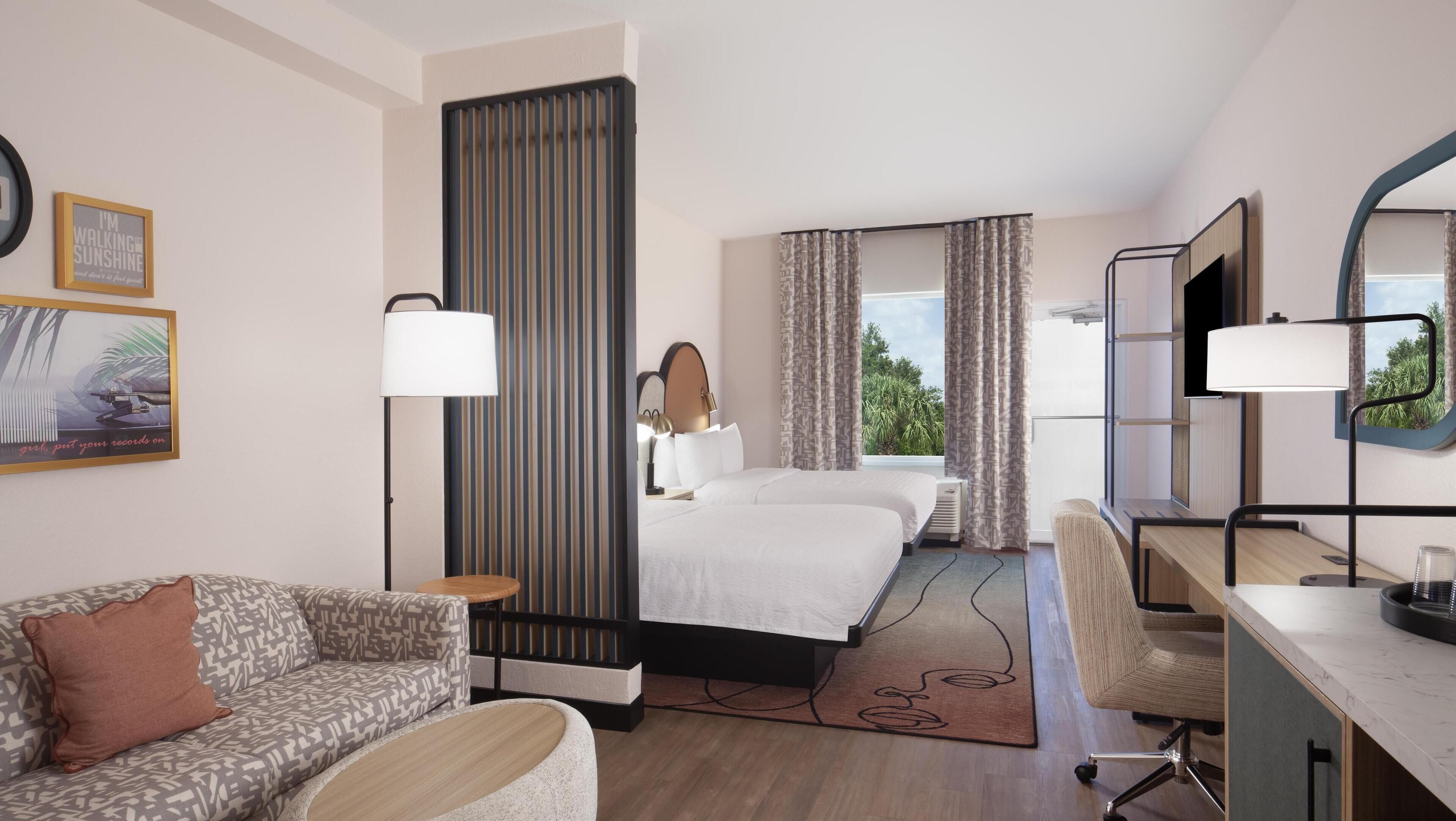Quarto de hotel com cama, escrivaninha, sofá e uma grande janela com cortinas.