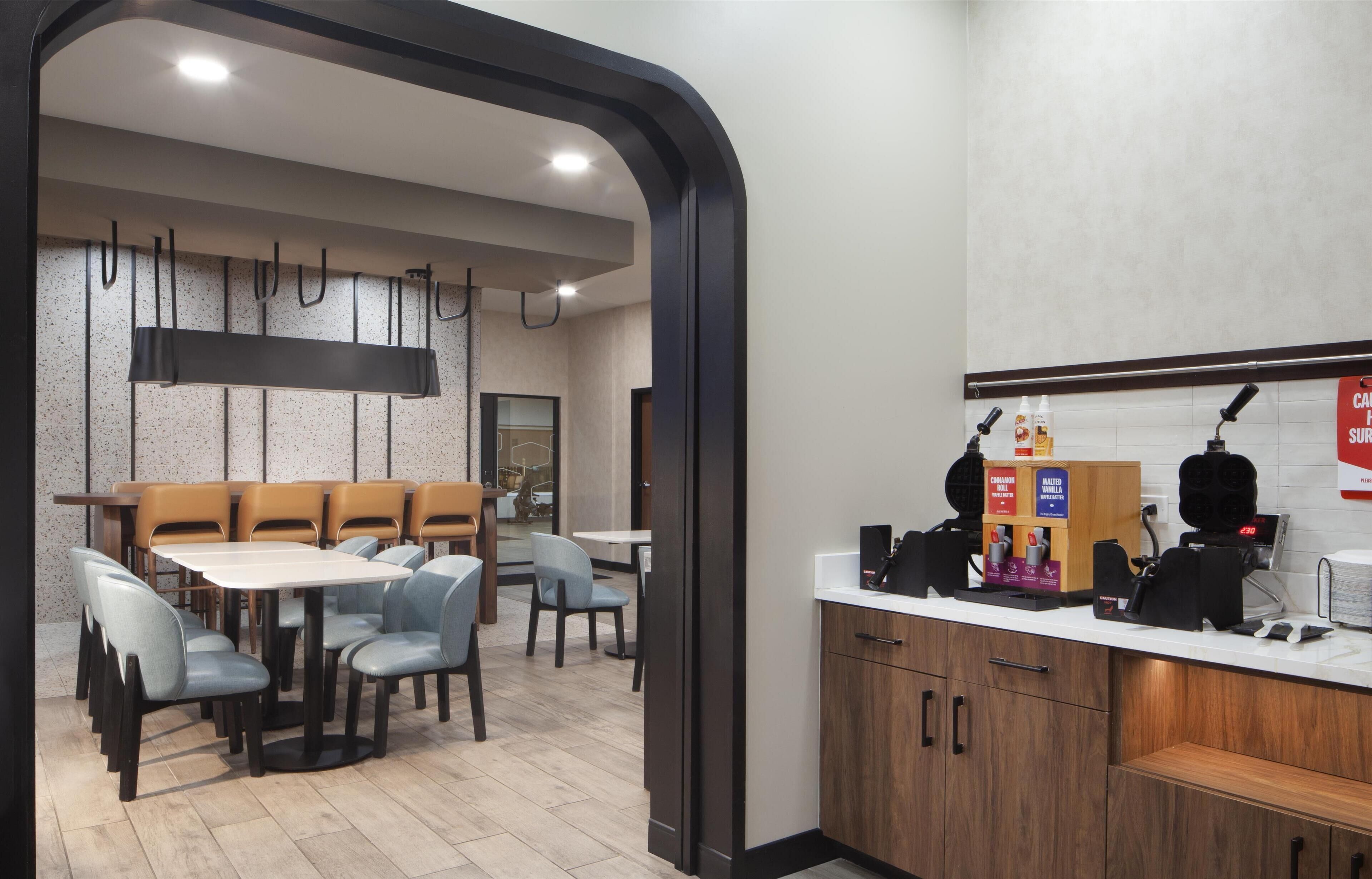 Un restaurante moderno con una larga mesa común y asientos individuales, una barra con máquina de café, y un cartel que anuncia una oferta especial.