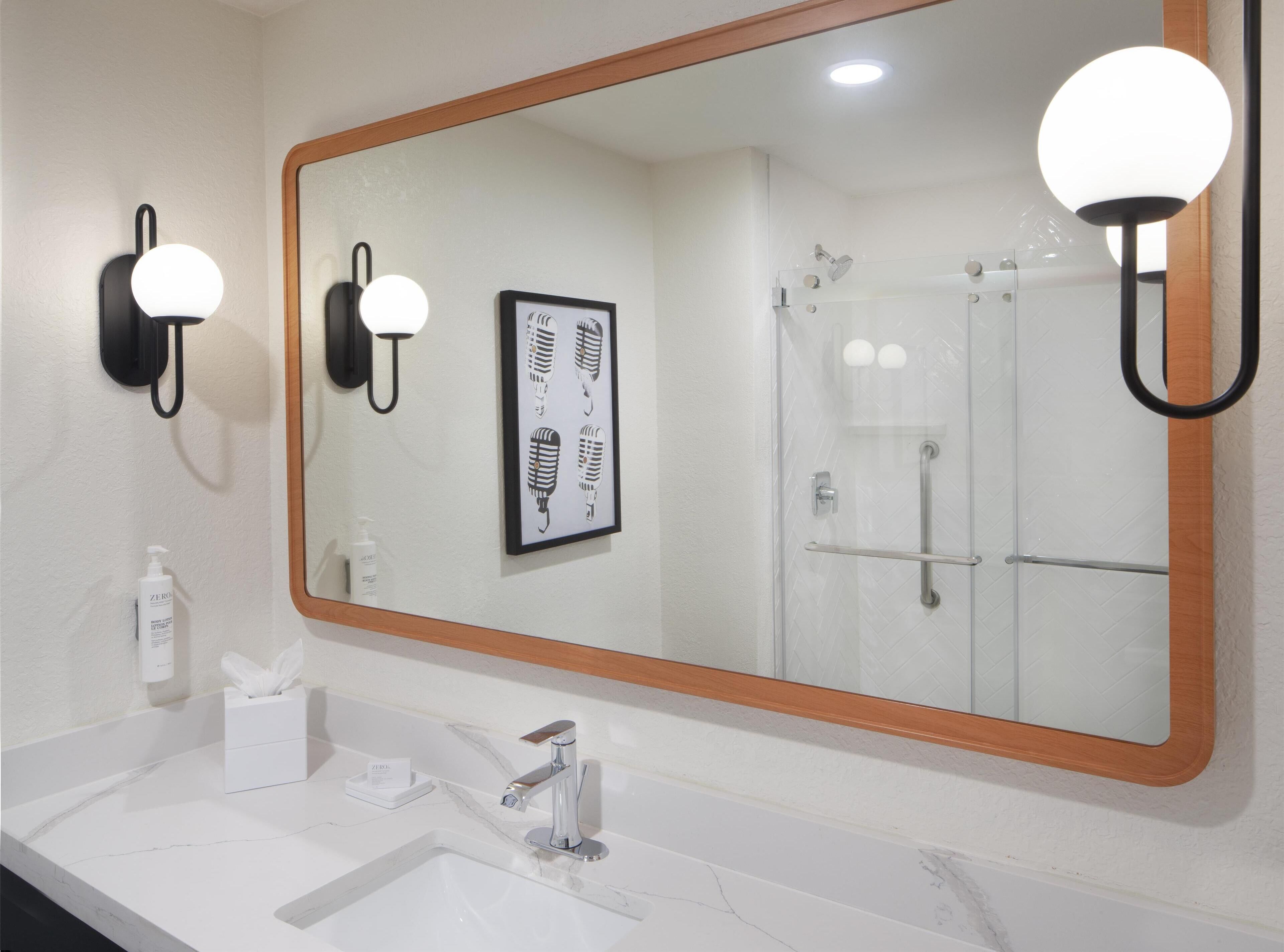 Un baño con un espejo grande, dos luces empotradas, un cuadro enmarcado, un lavamanos con encimera de mármol y una zona de ducha.