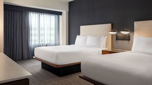 A hotel room with two beds, a desk, a large window with curtains, and wall-mounted lamps.