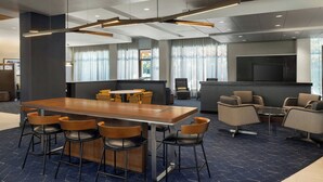 A modern hotel lobby with a long wooden table, bar stools, a seating area with sofas, a flat-screen TV, and large windows with curtains.