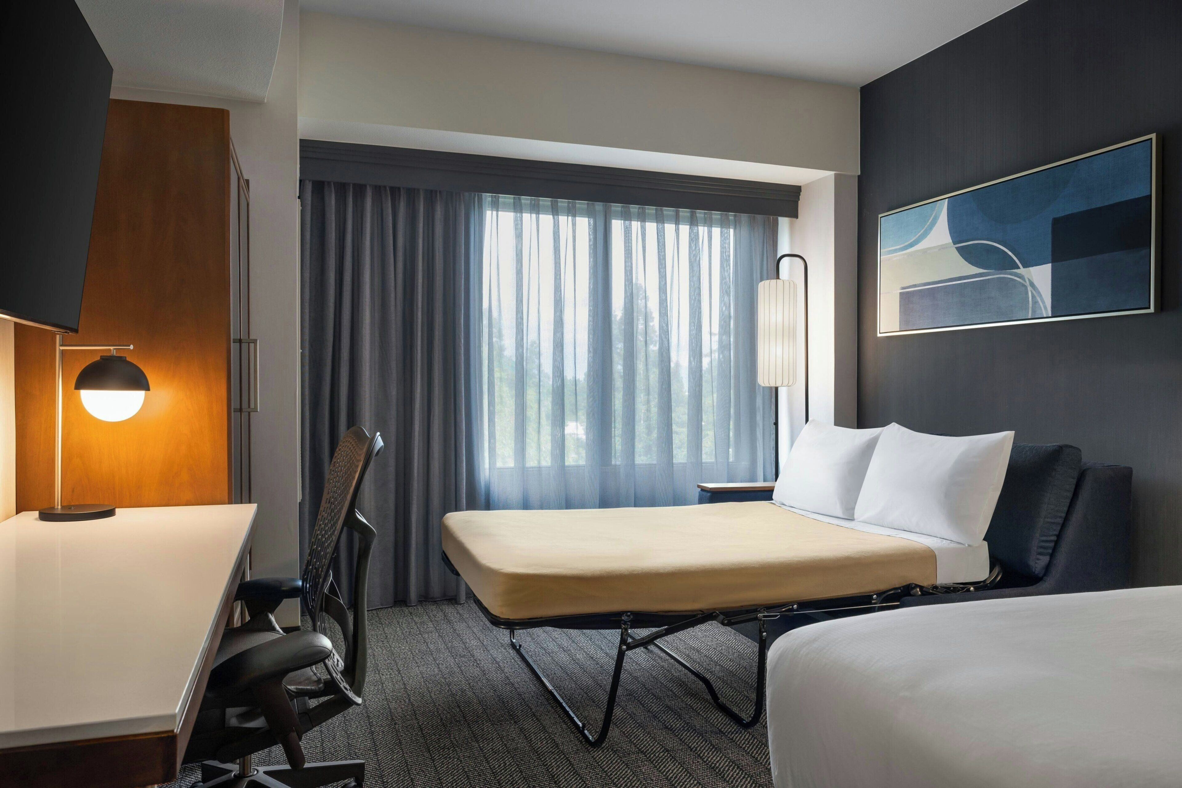 Una habitación de hotel con cama, escritorio, silla, lámpara, ventana con cortinas y un cuadro en la pared.