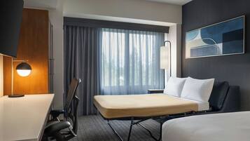 Una habitación de hotel con cama, escritorio, silla, lámpara, ventana con cortinas y un cuadro en la pared.