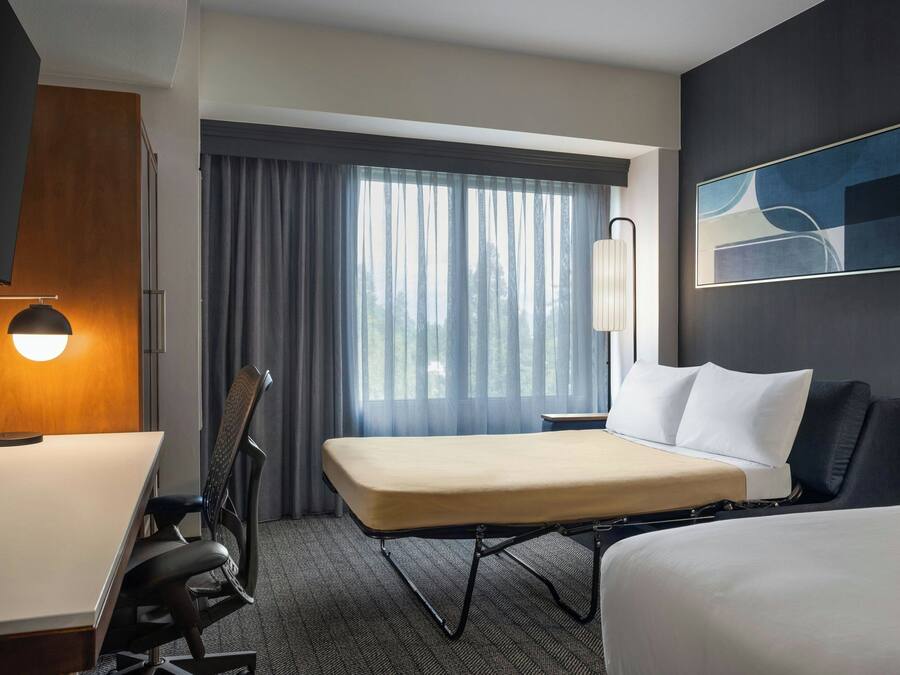 Una habitación de hotel con cama, escritorio, silla, lámpara, ventana con cortinas y un cuadro en la pared.