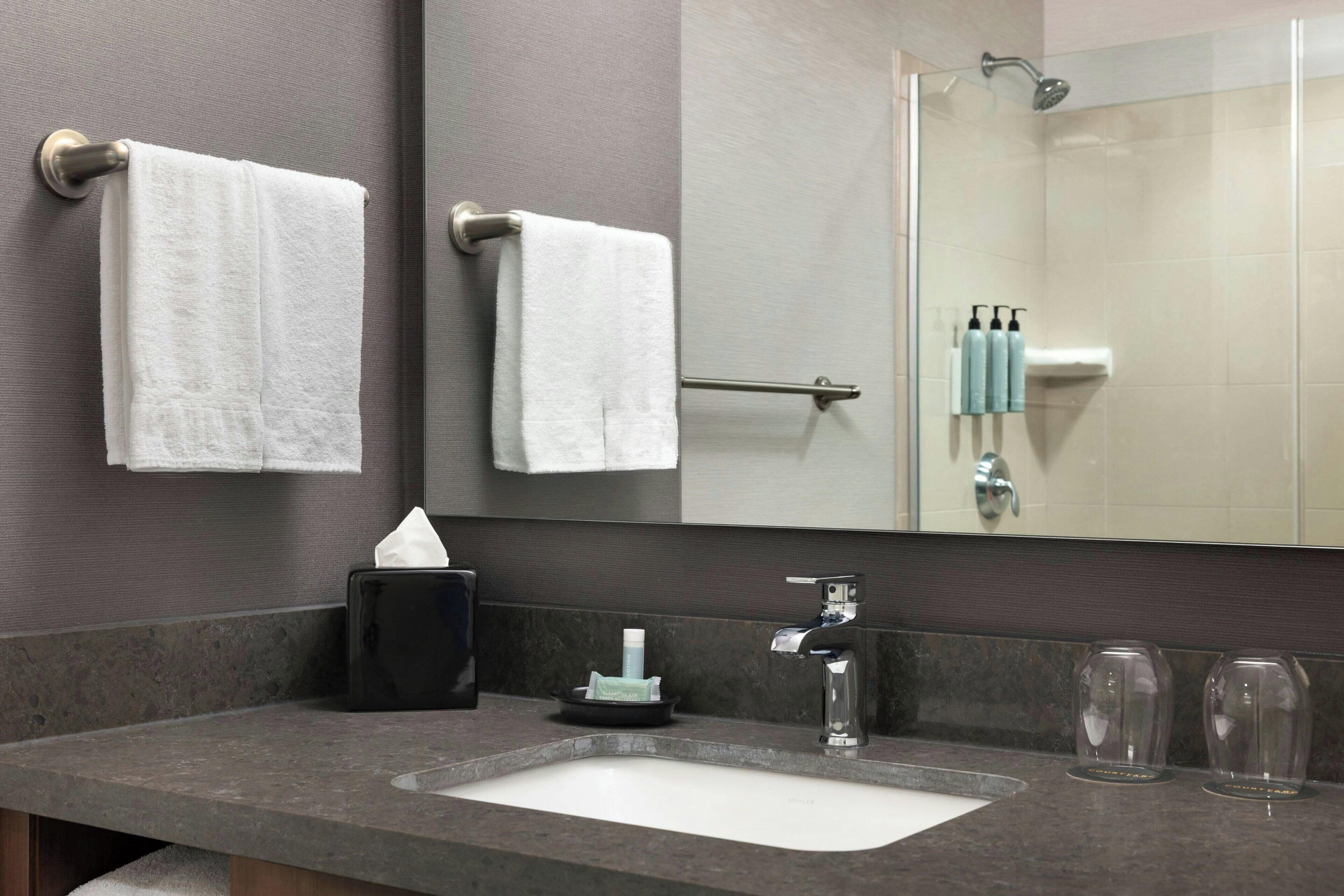 A bathroom with a sink, two towels, a mirror, a glass shower enclosure, and toiletries.