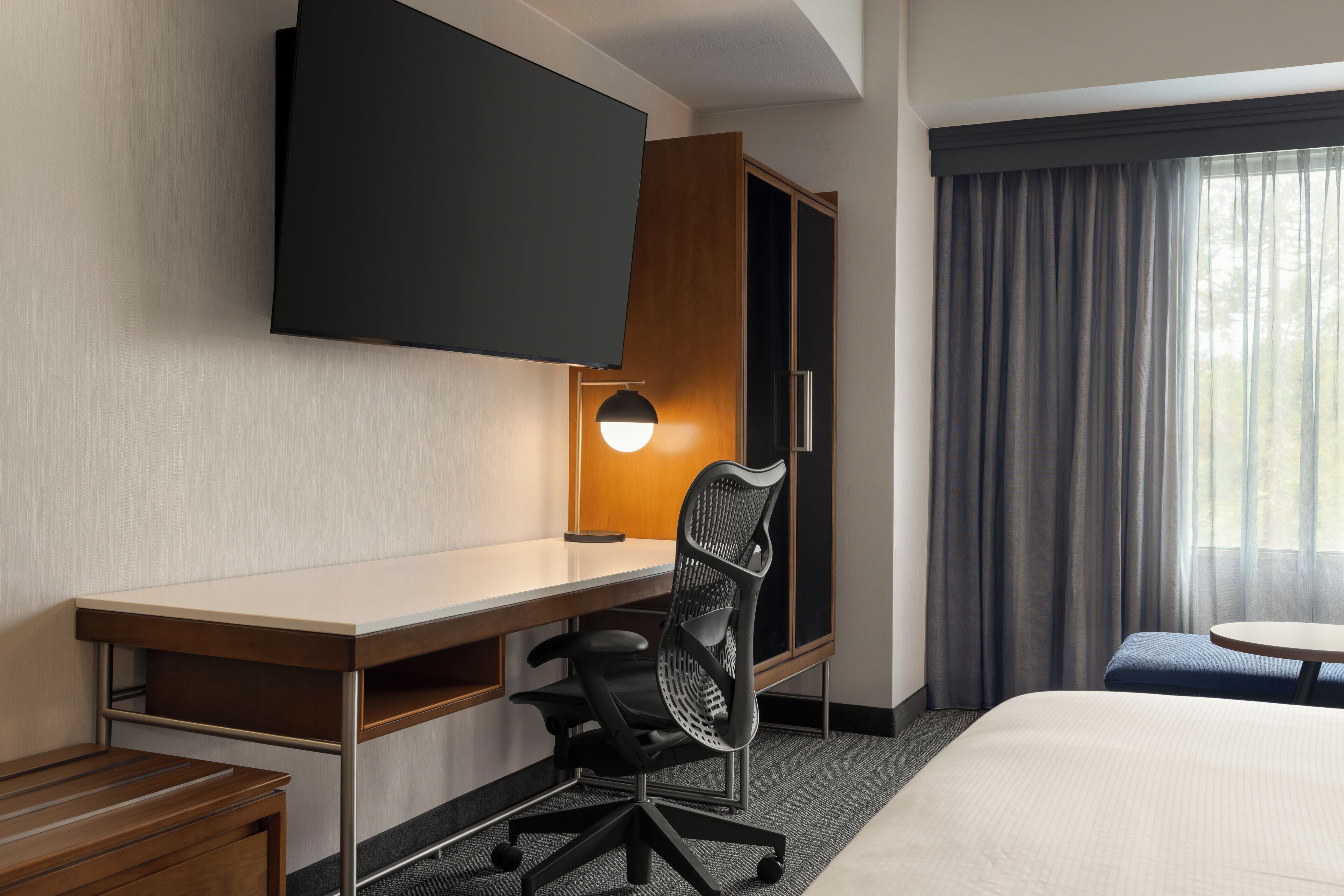 Habitación de hotel con escritorio, silla, cama, televisor y ventana con cortinas.