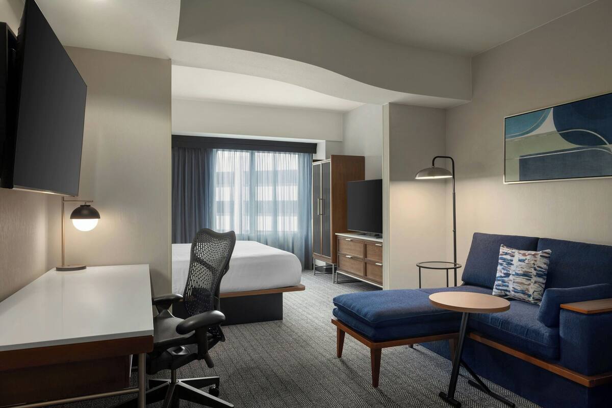 Una habitación de hotel con cama, escritorio, silla, sofá, televisión y lámpara.