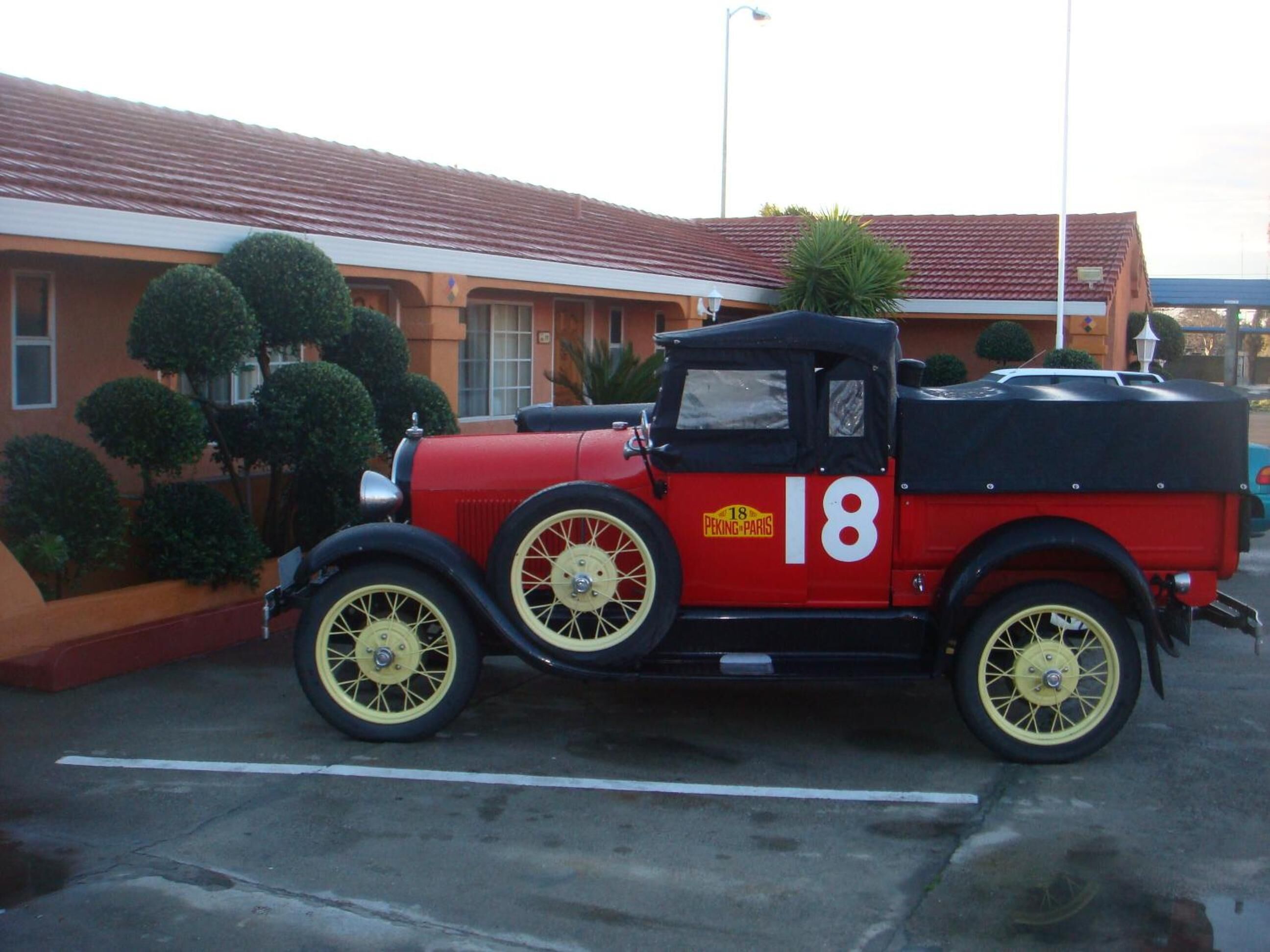 A vintage red and black car with the number 18 parked in front of a building with a red roof and green bushes.