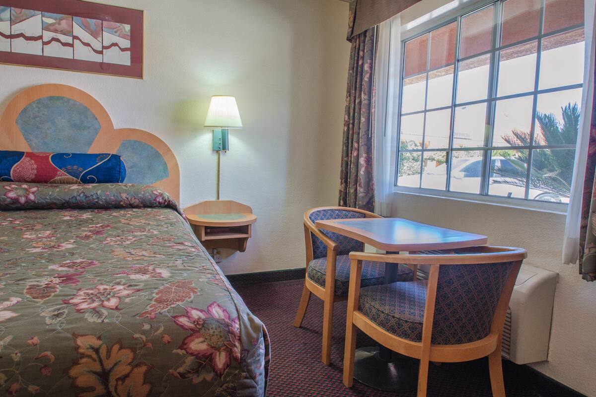A hotel room with a bed, a table, two chairs, a lamp, and a window with a view of the outside.