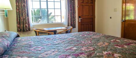 A bedroom with a floral bedspread, a window with curtains, a wooden door, and a small table.