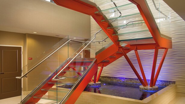 Una escalera moderna con barandillas de cristal y soportes estructurales rojos, que conduce a un nivel inferior con una zona de piscina decorativa.