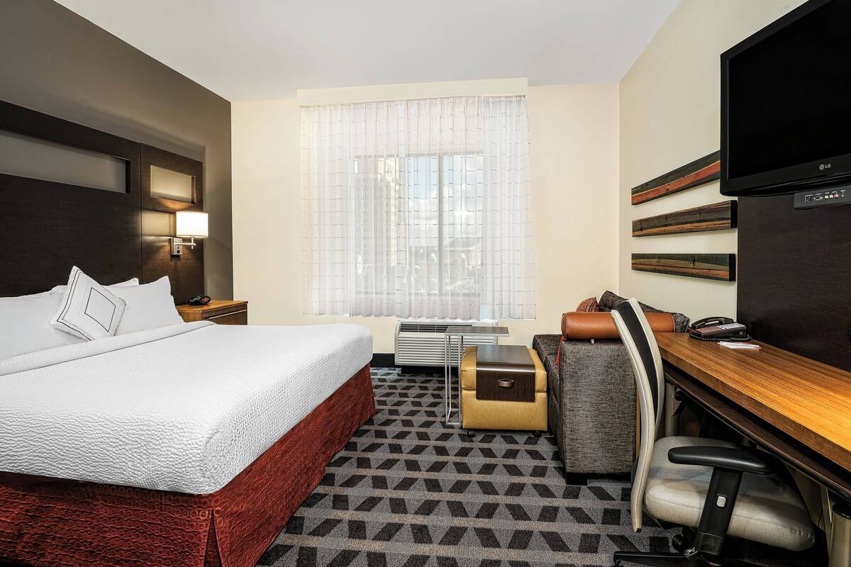 Ein Hotelzimmer mit einem großen Bett, einem Schreibtisch mit Stuhl, einem Flachbildfernseher, einem Fenster mit Vorhängen und einer Wandlampe.