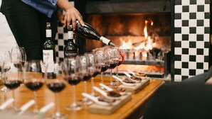 A person pouring wine into glasses on a table with a checkered wall and a fireplace in the background.