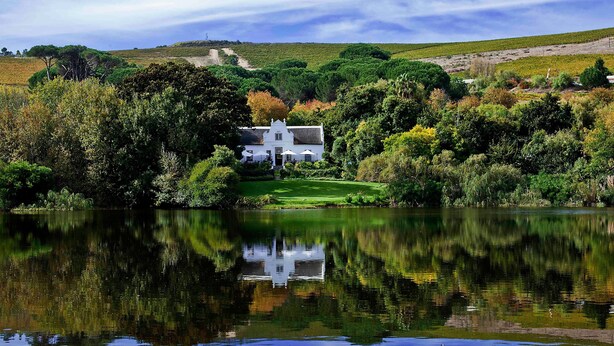 A white building with a thatched roof, surrounded by lush greenery and a calm lake reflecting the structure.