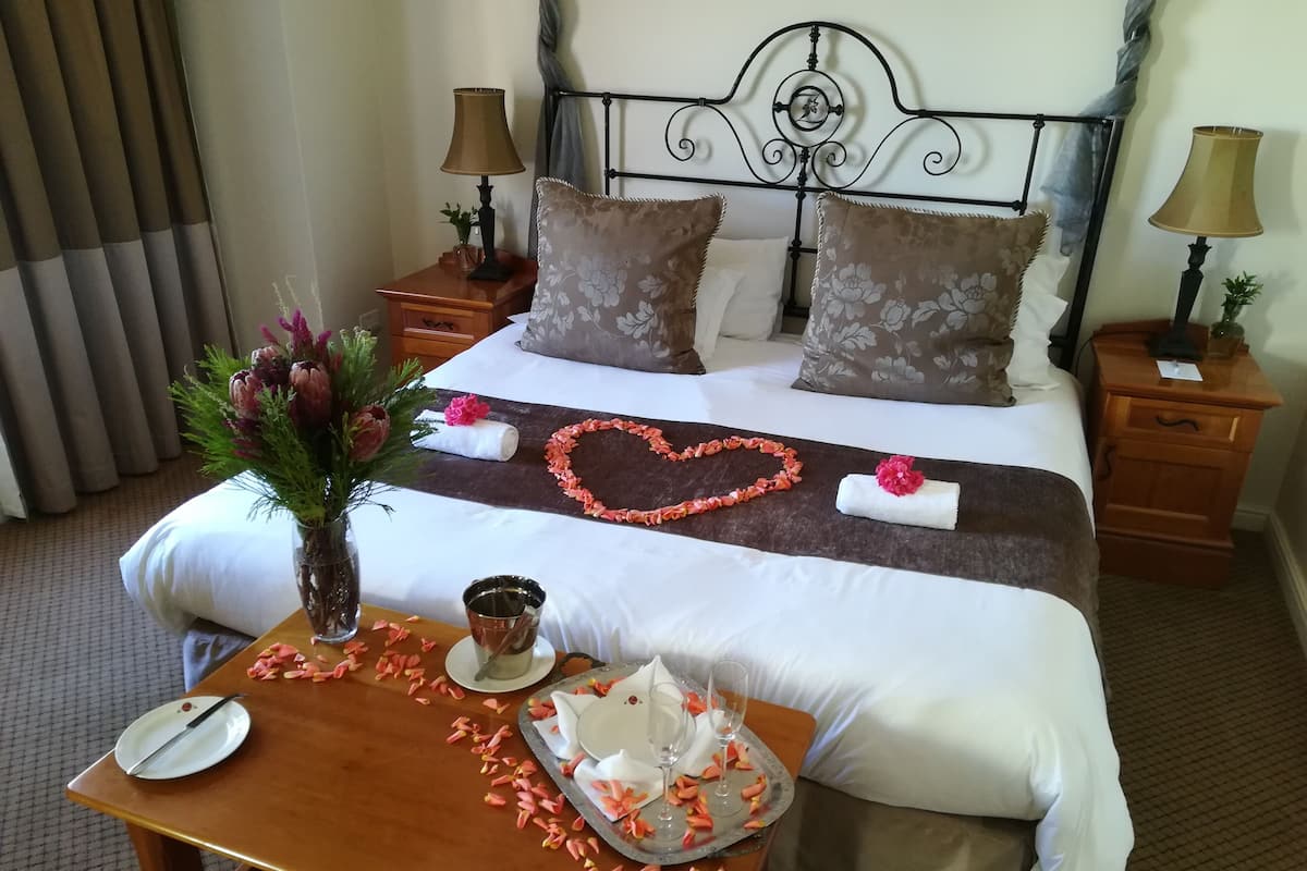 A neatly made bed with floral pillows, a heart-shaped decoration on the bedspread, and a tray with a cake and glasses on the side table.