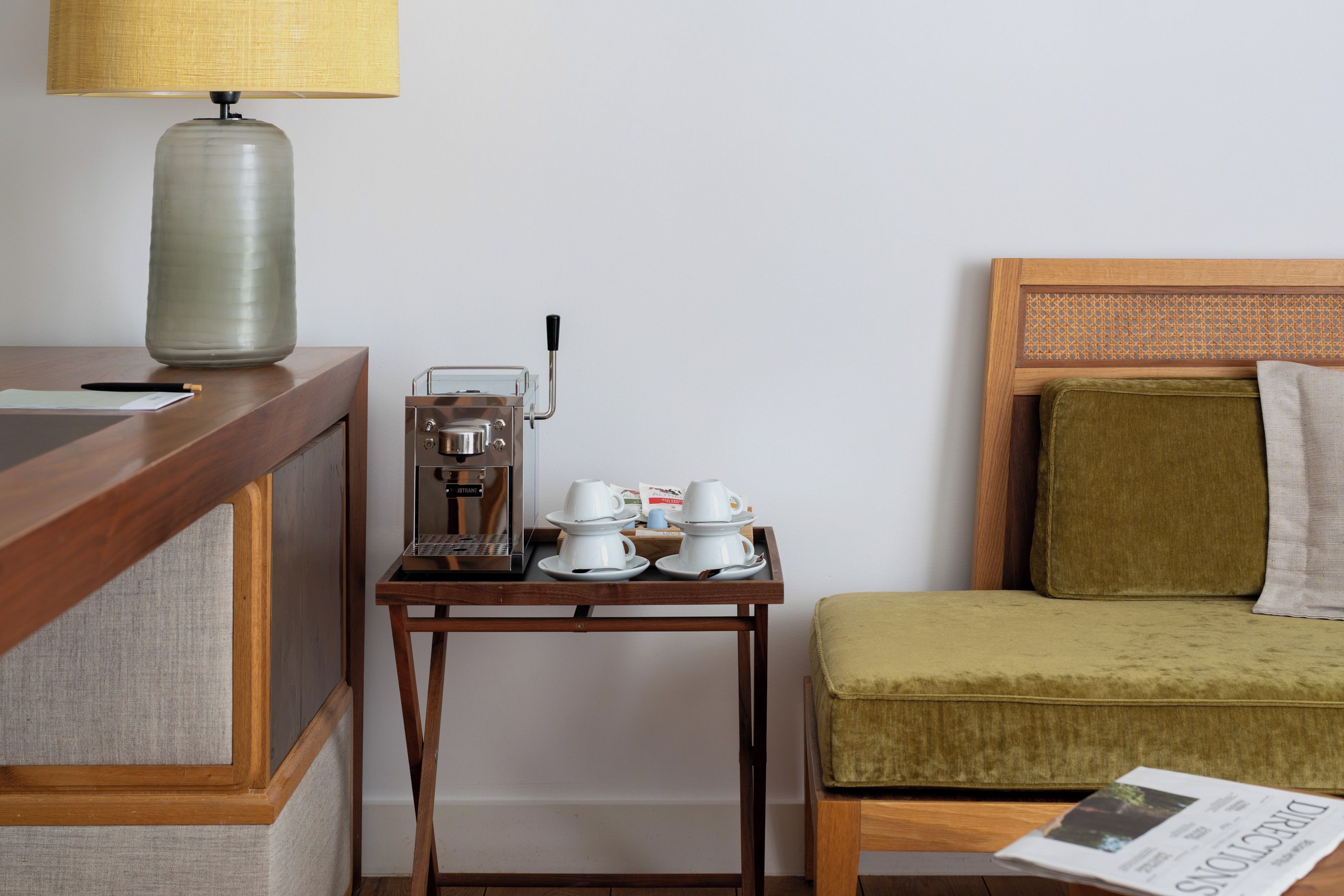 Ein Hotelzimmer mit einem Holztisch, einer Kaffeemaschine, einem kleinen Tisch mit Tassen und Untertassen, einem grün gepolsterten Stuhl und einer Zeitung auf dem Boden.