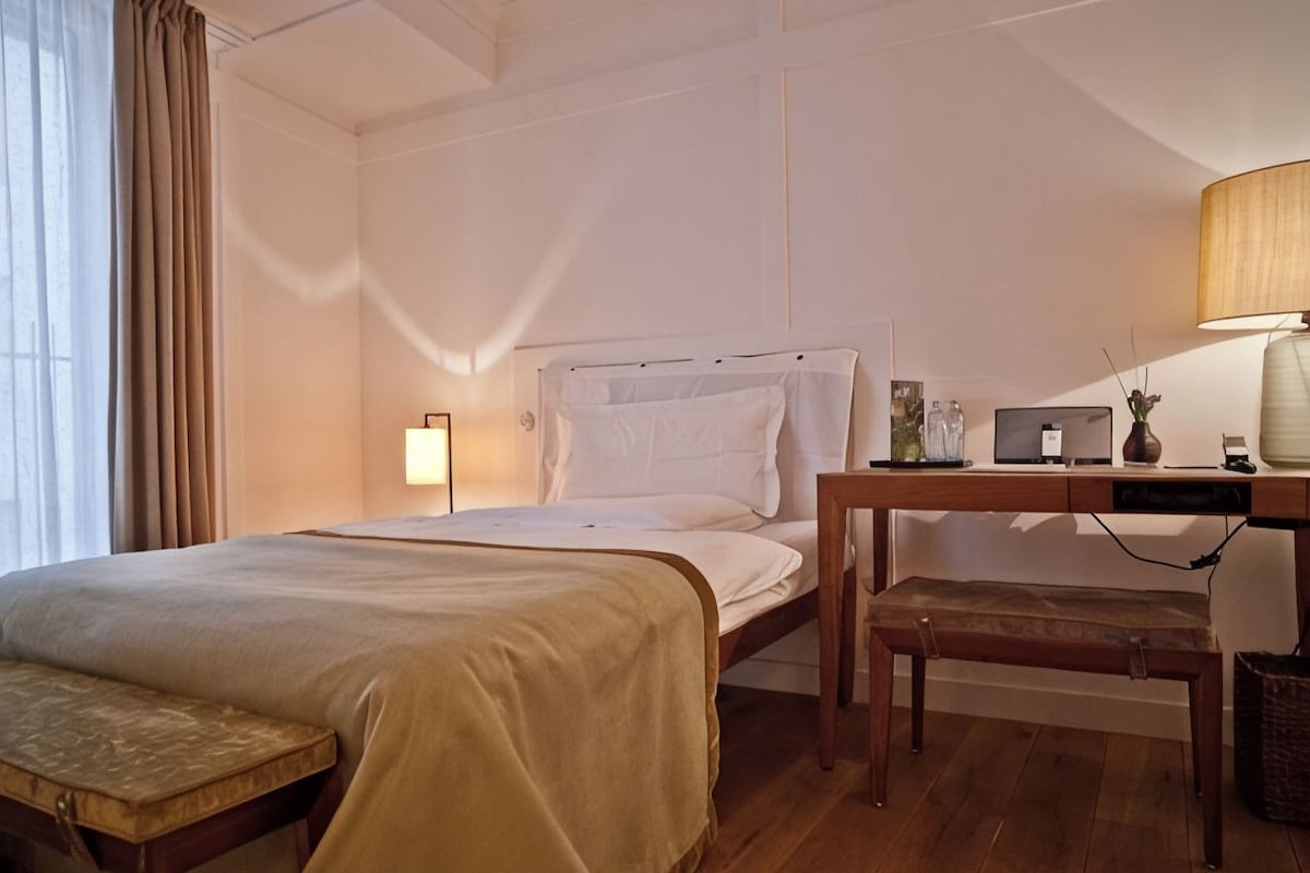 Quarto de hotel com cama, mesa de cabeceira, abajur e escrivaninha com cadeira.