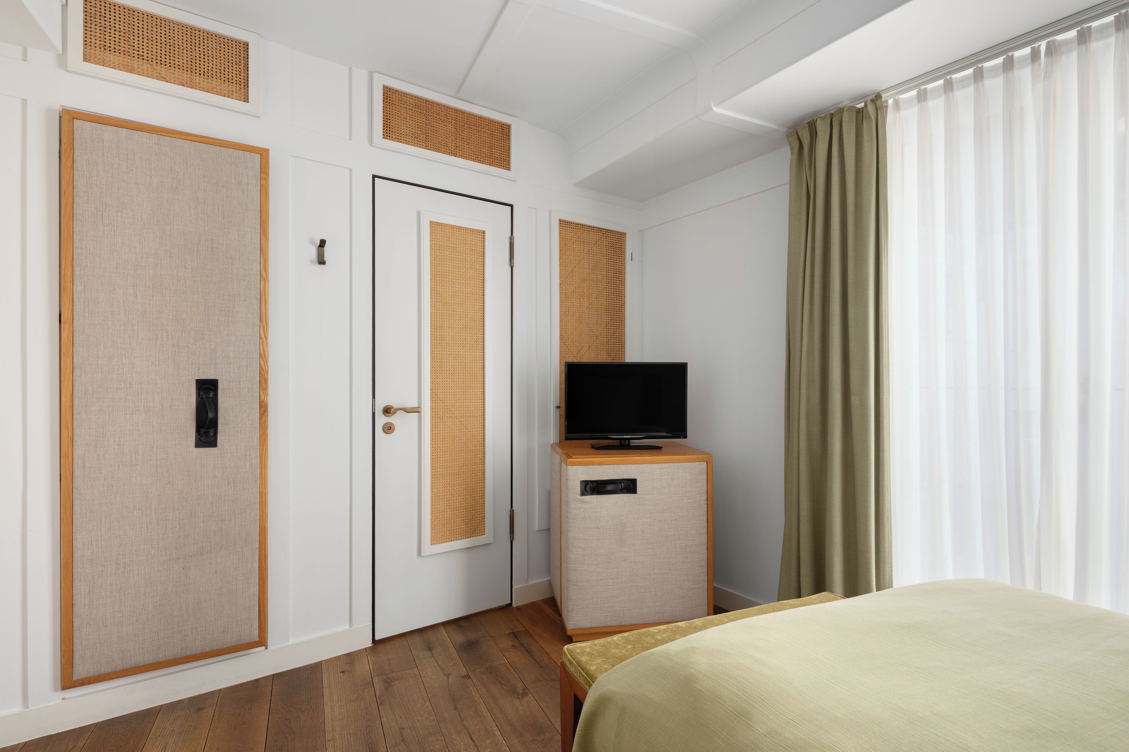 Habitación de hotel con cama, televisor sobre un mueble de madera, puerta con panel texturizado y ventana con cortinas.