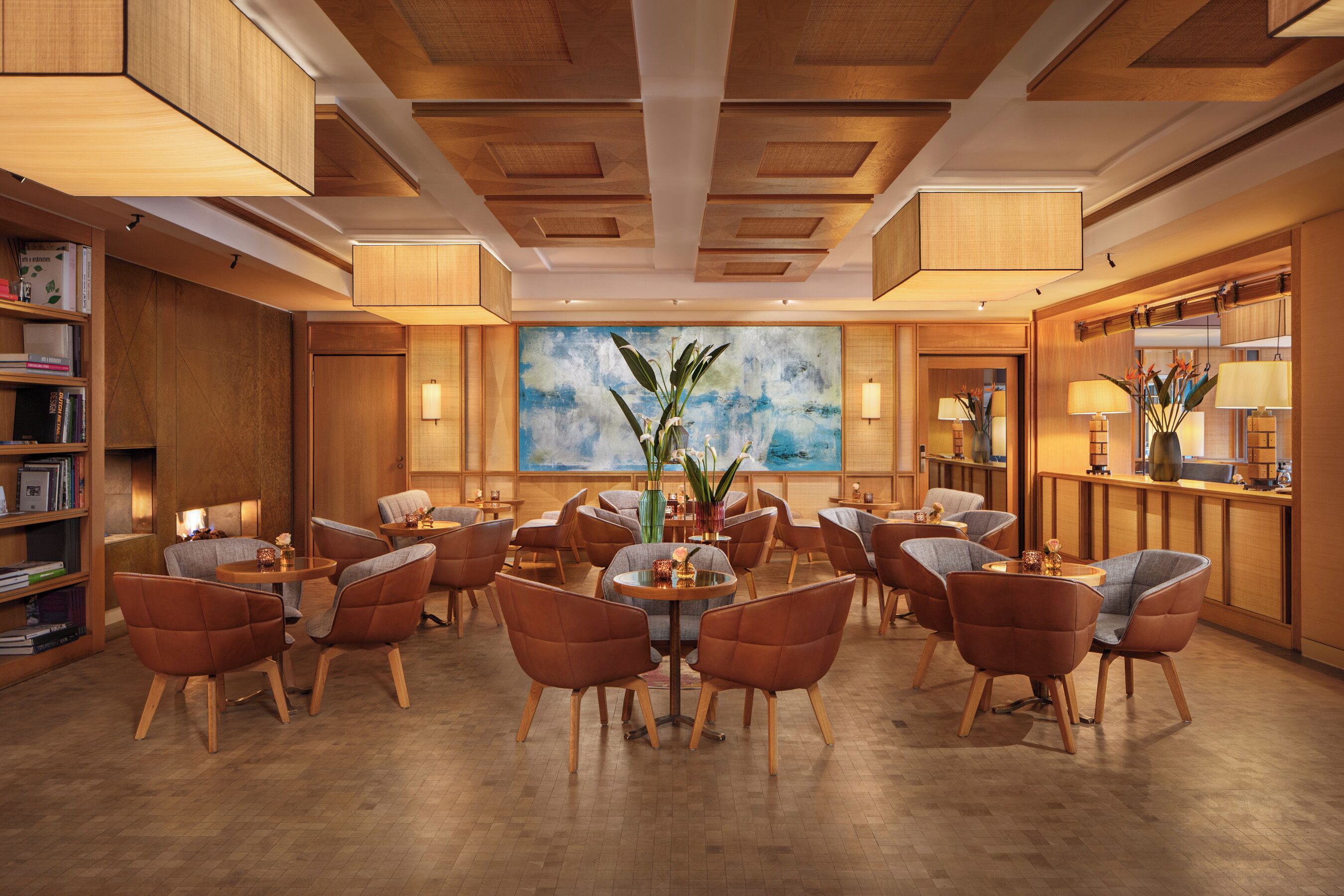 A spacious hotel lobby with wooden flooring, multiple seating areas, and a large abstract painting on the wall.